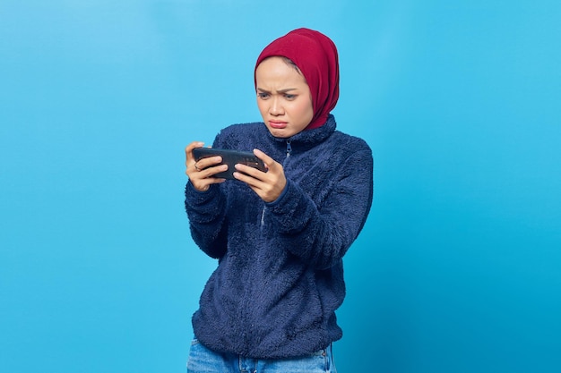 Shocked young asian woman playing a video game on a smartphone and looking up on blue background