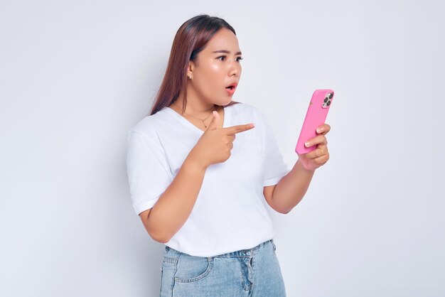 Шокированная молодая азиатка, указывающая пальцем на мобильный телефон, изолированный на белом фоне