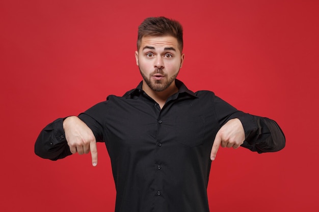 Shocked young bearded guy 20s in classic black shirt posing isolated on red wall background studio portrait. People sincere emotions lifestyle concept. Mock up copy space. Pointing index fingers down.