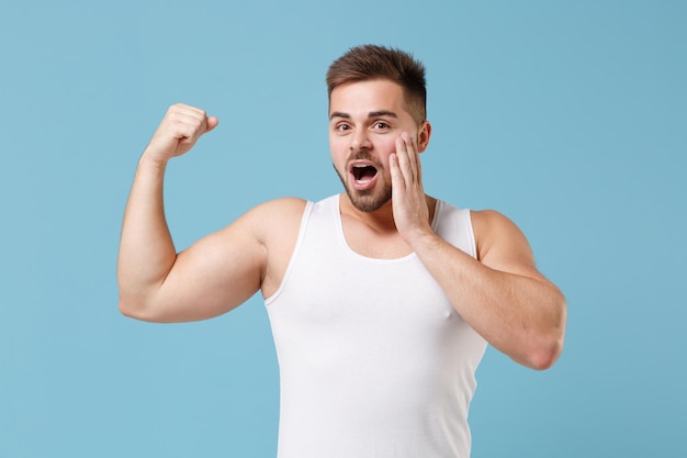 Shocked young bearded guy 20s in white singlet posing isolated on pastel blue wall background. Sport fitness healthy lifestyle concept. Mock up copy space. Showing biceps muscles, put hand on cheek.