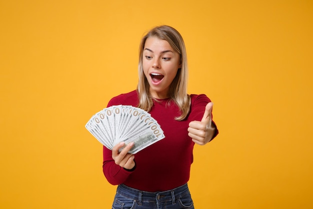 Shocked young blonde woman girl in casual clothes posing isolated on yellow orange background. people lifestyle concept. mock up copy space hold fan of cash money in dollar banknotes showing thumb up