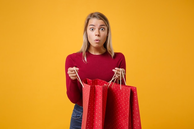 Photo shocked young blonde woman girl in casual clothes posing isolated on yellow orange background in studio. people lifestyle concept. mock up copy space. hold package bag with purchases after shopping.