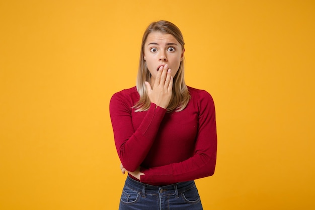 Shocked young blonde woman girl in casual clothes posing isolated on yellow orange background studio portrait. People sincere emotions lifestyle concept. Mock up copy space. Covering mouth with hand.