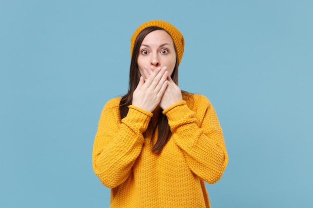 Shocked young brunette woman girl in sweater and hat posing isolated on blue wall background studio portrait. People sincere emotions lifestyle concept. Mock up copy space. Covering mouth with hands.