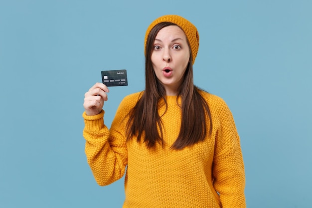 Shocked young brunette woman girl in yellow sweater and hat posing isolated on blue background studio portrait. People sincere emotions lifestyle concept. Mock up copy space. Hold credit bank card.