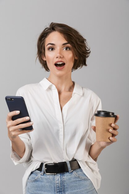 Shocked young business woman posing isolated over grey wall chatting by mobile phone drinking coffee