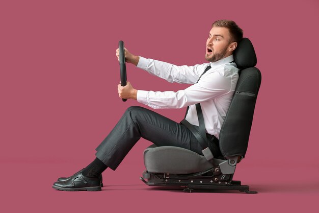 Shocked young businessman with steering wheel sitting on car seat against color background