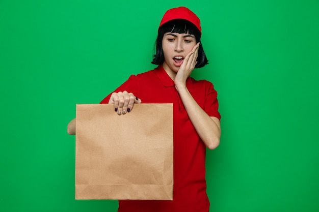 Shocked young caucasian delivery woman holding and looking at paper food bag