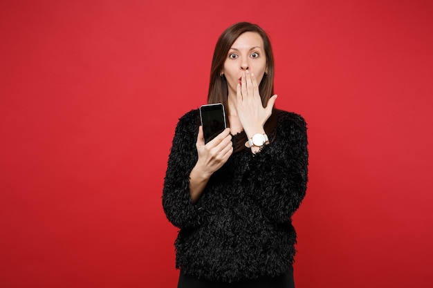 Shocked young girl in black fur sweater covering mouth with hand holding mobile phone with blank empty screen isolated on red background. People sincere emotions lifestyle concept. Mock up copy space.
