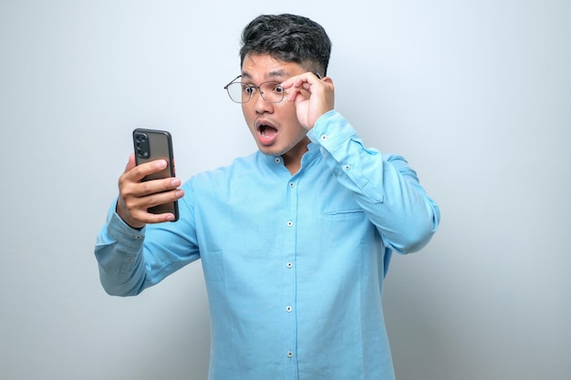 Шокированный молодой симпатичный азиат с помощью смартфона читает хорошие новости онлайн изолированно