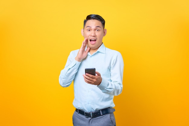 Shocked young handsome businessman holding smartphone and looking camera on yellow background