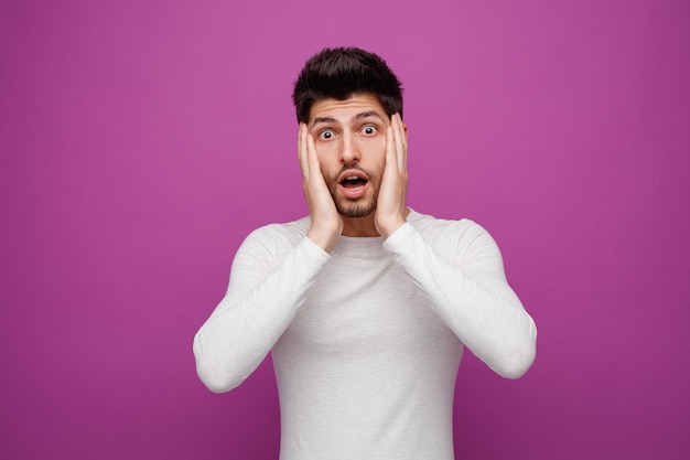 Shocked young handsome man keeping hands on face looking at camera on purple background