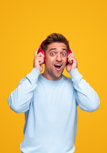 Shocked young man in blue sweatshirt adjusting headphones and looking at camera with opened mouth while listening to loud music against yellow background