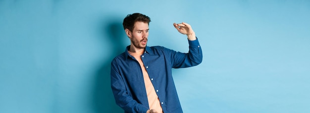 Shocked young man looking at empty space and showing big size object standing on blue background