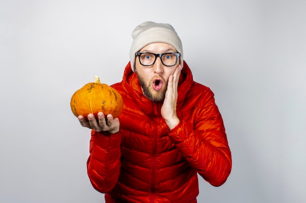Shocked young man in a red jacket, hat, holds a pumpkin and with his hand hold on his cheek
