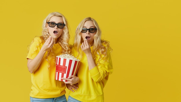 Shocked young models women in 3d glasses eating popcorn, looks scary Movie. Isolated on yellow wall