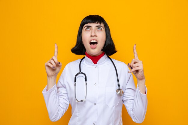 Shocked young pretty caucasian girl in doctor uniform with stethoscope looking and pointing up pointing