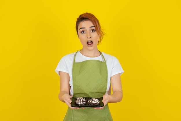 Shocked young redhead holding fresh cookies on yellow wall 