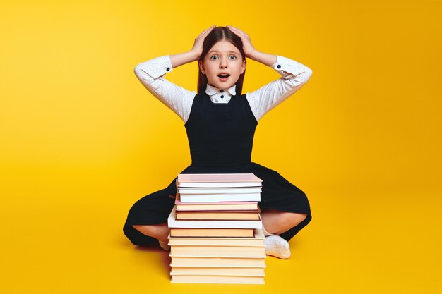 Foto ragazza scolaressa scioccata in uniforme che tiene le mani sulla testa mentre è seduta vicino a una pila di libri