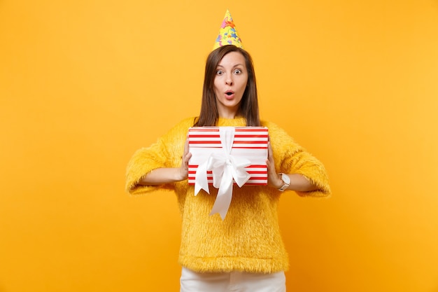 Shocked young woman in birthday hat holding red box with gift present celebrating, enjoying holiday isolated on bright yellow background. People sincere emotions, lifestyle concept. Advertising area.