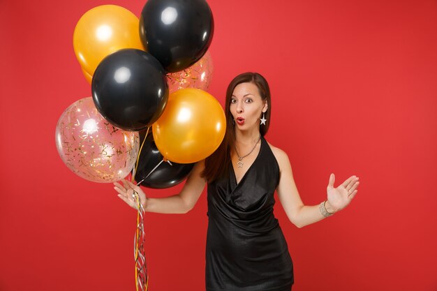 Shocked young woman in black dress celebrating spreading hands holding air balloons isolated on bright red background. International Women's Day, Happy New Year, birthday mockup holiday party concept.