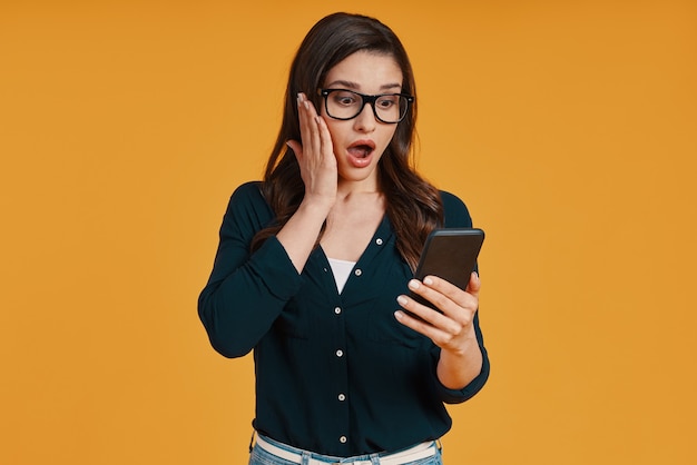 Shocked young woman in casual clothing using smart phone while standing against yellow background