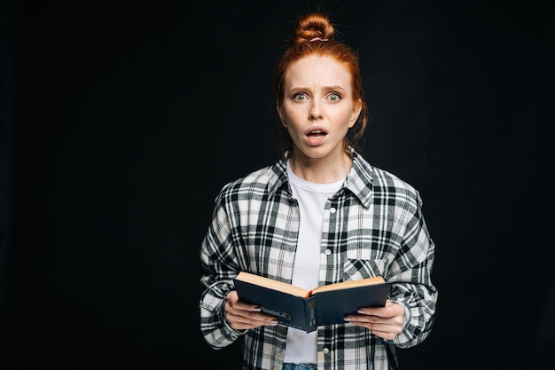 Shocked young woman college student with opened mouth holding books and looking at camera on black isolated background Pretty redhead lady model emotionally showing facial expressions in studio