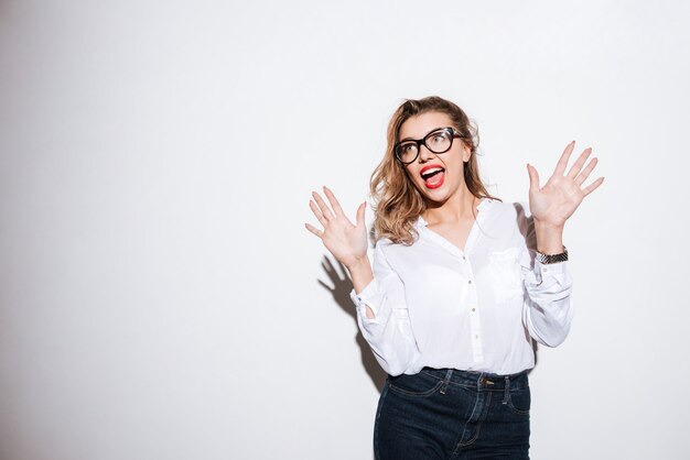 Shocked young woman in eyeglasses with raised hands and opened mouth over white wall