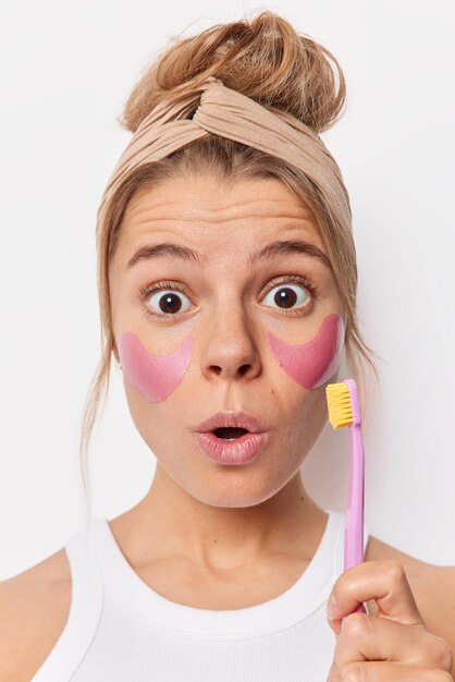 Shocked young woman feels amazed applies beauty patches holds toothrush undergoes daily hygiene treatments and routines during morning wears headband holds breath isolated over white background.