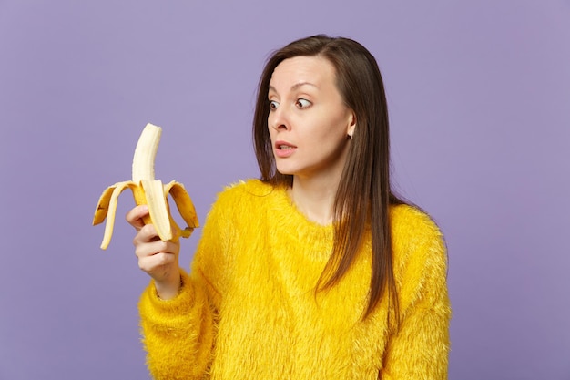 Shocked young woman in fur sweater holding in hand, looking on fresh ripe banana fruit isolated on violet pastel wall background. People vivid lifestyle, relax vacation concept. Mock up copy space.