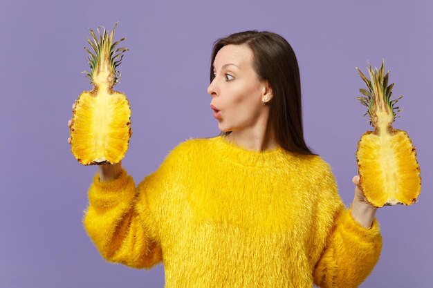 Shocked young woman in fur sweater looking aside holding halfs of fresh ripe pineapple fruit isolated on violet pastel background. People vivid lifestyle, relax vacation concept. Mock up copy space.