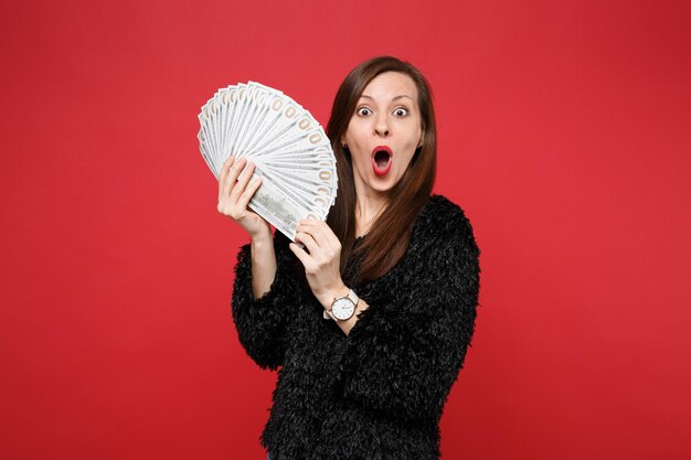 Shocked young woman in fur sweater with opened mouth holding fan of money in dollar banknotes, cash money isolated on red background. People sincere emotions, lifestyle concept. Mock up copy space.