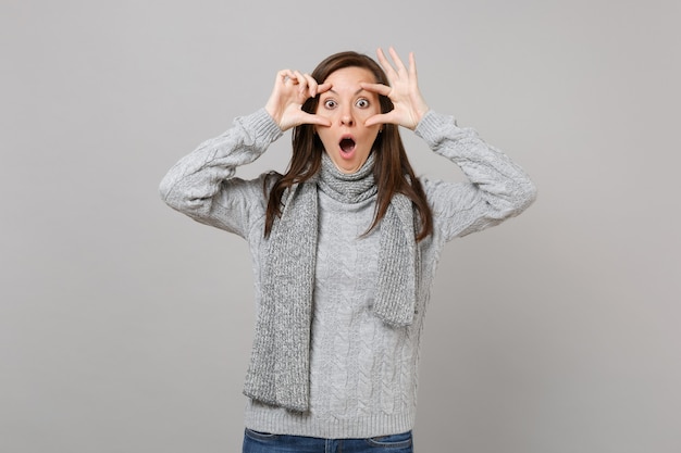 Shocked young woman in gray sweater, scarf keeping mouth wide open, stretching eyelids isolated on grey background. Healthy fashion lifestyle, people emotions, cold season concept. Mock up copy space.