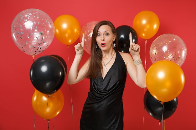 Scioccata giovane donna in abitino nero che celebra puntando il dito indice su palloncini d'aria di sfondo rosso brillante. giornata internazionale della donna, felice anno nuovo, concetto di festa di compleanno mockup.