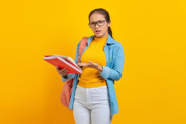 Shocked young woman student in denim clothes with backpack\
pointing notebook with hand isolated on yellow background education\
in high school university college concept