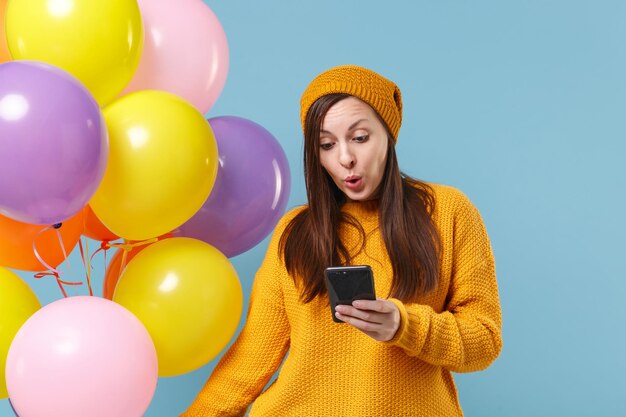 青い背景に分離されたセーターの帽子のポーズでショックを受けた若い女性。誕生日の休日のパーティー、人々の感情の概念。コピースペースをモックアップします。携帯電話を使ってカラフルな風船を持ってお祝いしましょう。