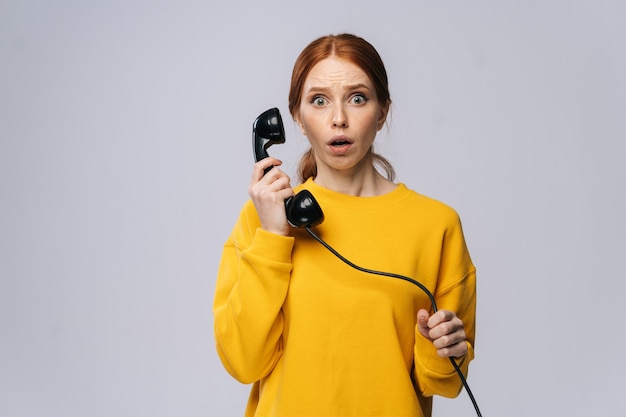 Shocked young woman wearing stylish yellow sweater talking on retro phone looking at camera