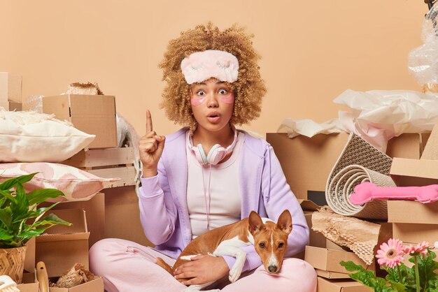 Shocked young woman wears domestic clothes points index finger\
holds breath from amazement shows something astonishing poses with\
pedigree dog surrounded by cardboard boxes full of belongings