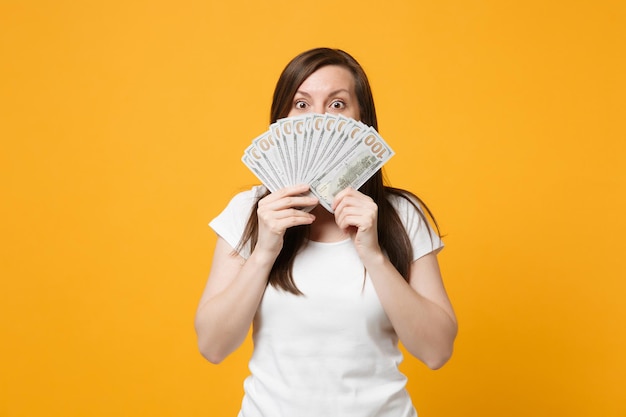 shocked young woman in white casual clothes covering face with fan of cash money in dollar banknotes isolated on yellow orange wall background in studio. people lifestyle concept. mock up copy space.
