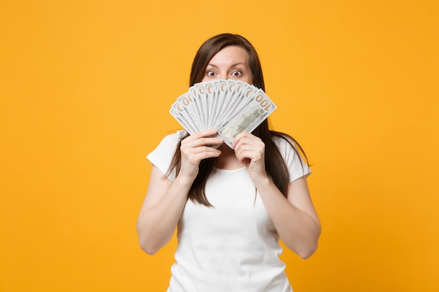 Shocked young woman in white casual clothes covering face with fan of cash money in dollar banknotes isolated on yellow orange wall background in studio. people lifestyle concept. mock up copy space.