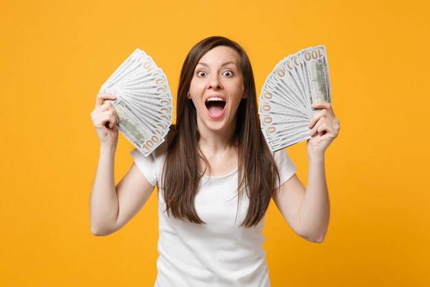 shocked young woman in white clothes keeping mouth wide open holding fan of cash money in dollar banknotes isolated on yellow orange background in studio. people lifestyle concept. mock up copy space.
