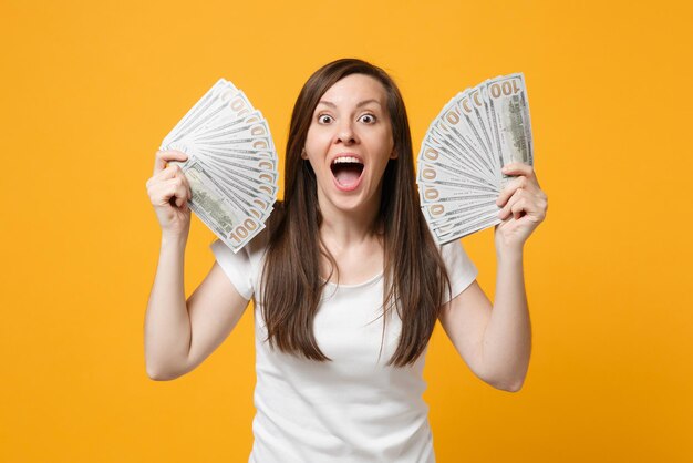 Shocked young woman in white clothes keeping mouth wide open holding fan of cash money in dollar banknotes isolated on yellow orange background in studio. People lifestyle concept. Mock up copy space.