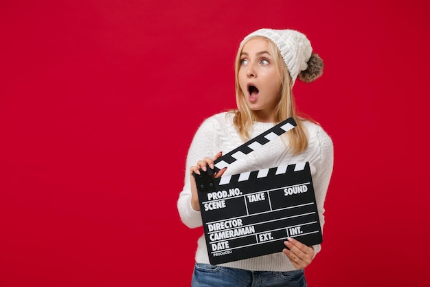 Shocked young woman in white sweater, hat isolated on red background. Healthy fashion lifestyle, cold season concept. Mock up copy space. Hold classic black film making clapperboard, looking aside.