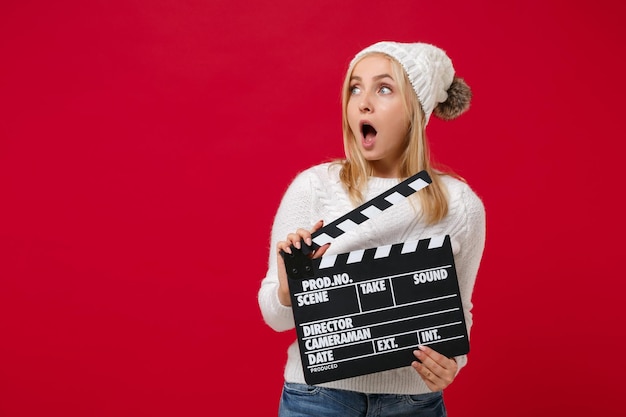 Giovane donna scioccata in maglione bianco, cappello isolato su sfondo rosso. stile di vita sano della moda, concetto di stagione fredda. simula lo spazio della copia. tieni il classico ciak per la produzione di film neri, guardando da parte.