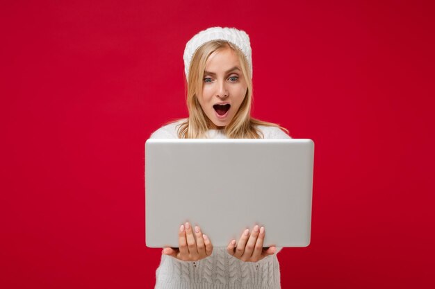 Shocked young woman in white sweater hat isolated on red wall background in studio. Healthy fashion lifestyle people sincere emotions, cold season concept. Mock up copy space. Hold laptop pc computer.