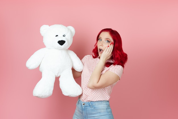 Шокированная молодая женщина с открытым ртом и рыжими волосами держит большого белого плюшевого мишку