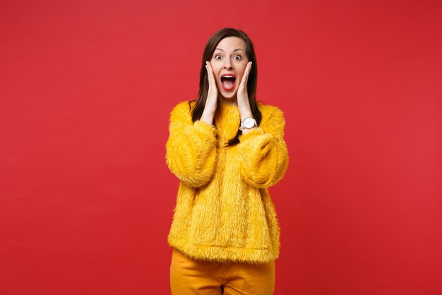 Shocked young woman in yellow fur sweater keeping mouth wide open, putting hands on cheeks isolated on bright red background in studio. People sincere emotions, lifestyle concept. Mock up copy space.