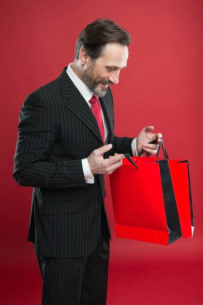 충격적인 할인. 성숙한 사업가 보유 종이 가방 선물 빨간색 배경. 쇼핑 개념입니다. 크리스마스 선물. 즐거운 휴일 보내세요. 남자의 행복한 얼굴은 새해를 축하합니다. 발렌타인 데이 선물. 좋은 구매입니다.