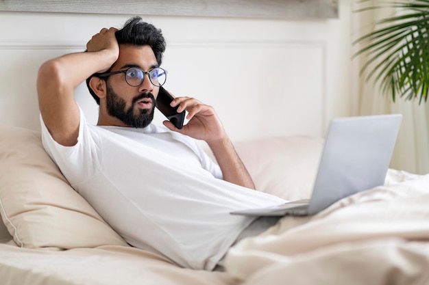 Шокирующие новости эмоциональный мужчина разговаривает по мобильному и использует ноутбук в постели