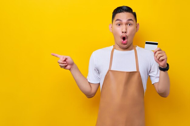 Shokced jonge Aziatische man 20s barista-medewerker in een bruin schort werkend in de coffeeshop met creditcard wijzende vinger opzij op gele achtergrond Klein bedrijf opstarten concept